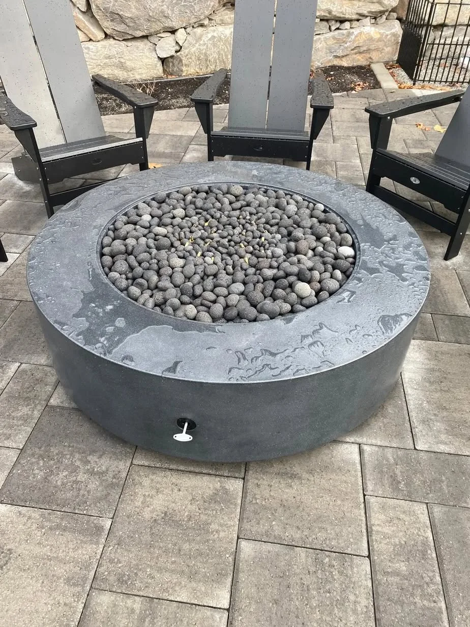 Round gas fire pit filled with lava rock on a paver patio beside a boulder wall